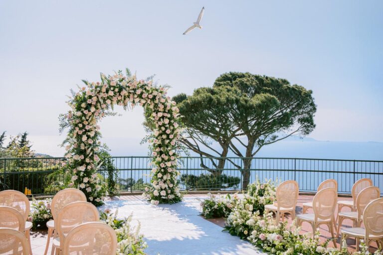 A rose and greenery arch on a white terrace above the sea, cane chairs either side of a white aisle and an umbrella pine beside it, Weddings Amalfi