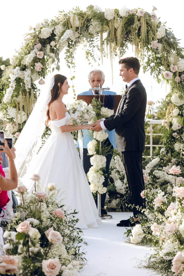 The vows under an arch of white roses and greenery, the celebrant reading, Weddings Amalfi
