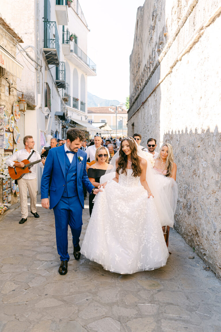 The couple walking a narrow stone lane behind a guitarist, guests following, Weddings Amalfi