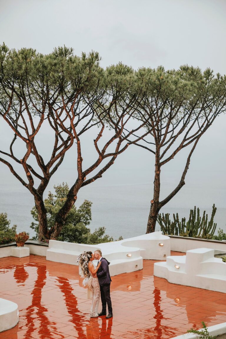 Two umbrella pines over a terracotta terrace above the sea, the couple small beneath them, Weddings Amalfi