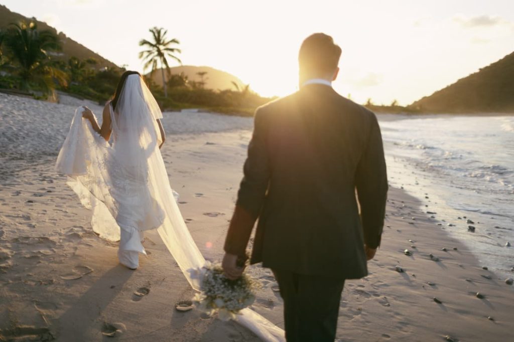 A bride and groom walking along the shore at sunset, the veil trailing behind