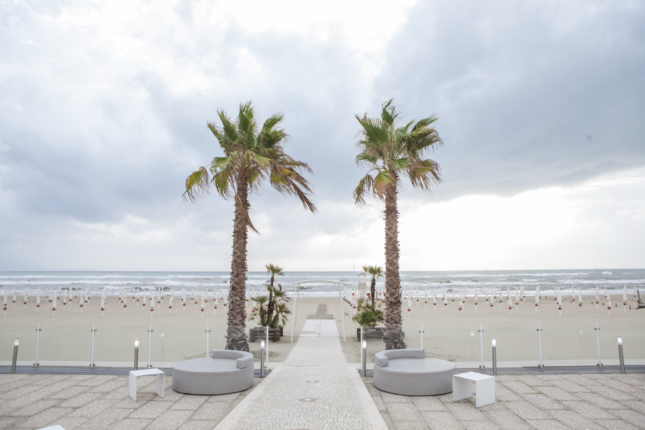 A ceremony aisle laid across a seafront terrace to the beach, two palms framing the view of the sea, WEM Eventi