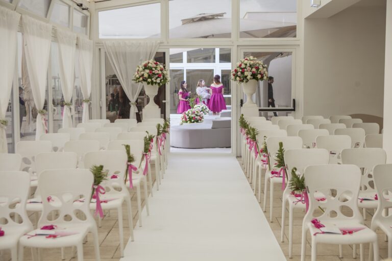 Rows of white chairs dressed with pink ribbon either side of a white aisle in a glazed seaside veranda, WEM Eventi
