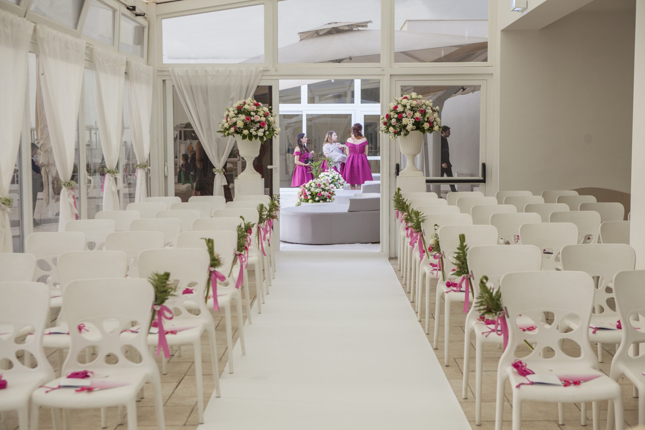 Rows of white chairs dressed with pink ribbon either side of a white aisle in a glazed seaside veranda, WEM Eventi