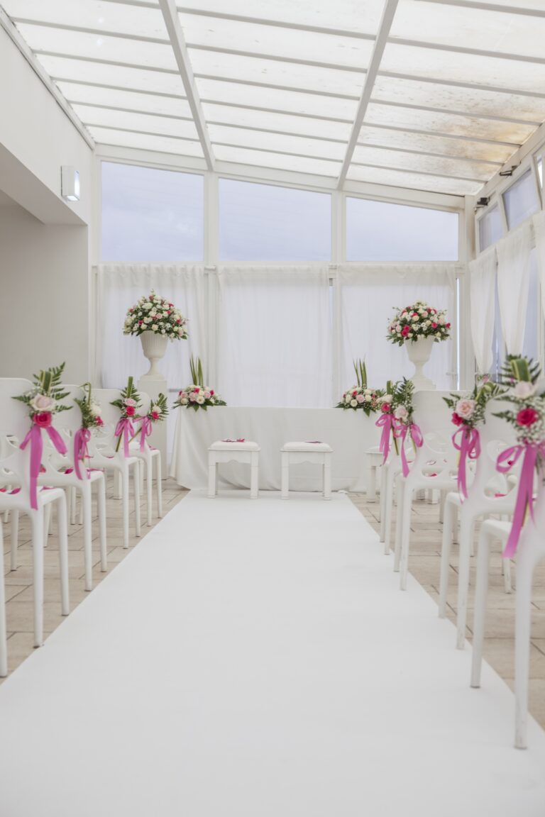 A white carpeted aisle leading to the ceremony table under a glass roof, chairs tied with pink ribbon, WEM Eventi
