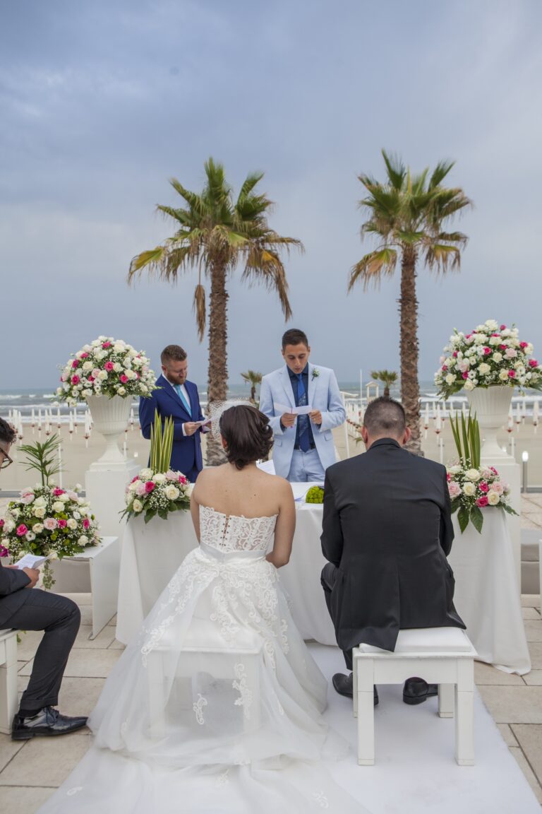 A ceremony by the sea, the couple seated at the ceremony table between urns of roses and two palms, WEM Eventi