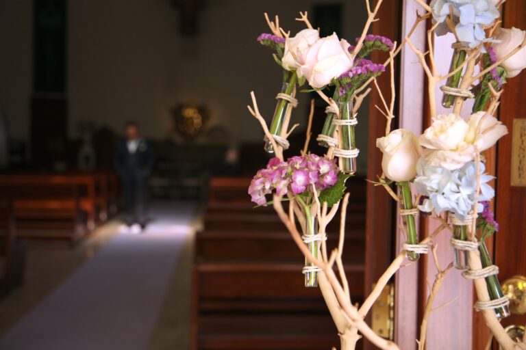 Roses, hydrangea and sweet william in glass vials on white branches at the end of a church pew, looking down the aisle to the altar, WEM Eventi