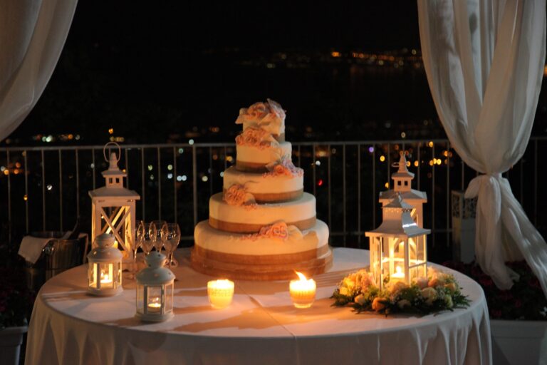 A tiered wedding cake on a candlelit terrace at night, white lanterns either side and city lights below, WEM Eventi