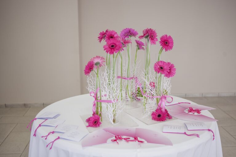 Pink gerberas in slim glass vases on white branches, set on a round table with fan-shaped cards, WEM Eventi