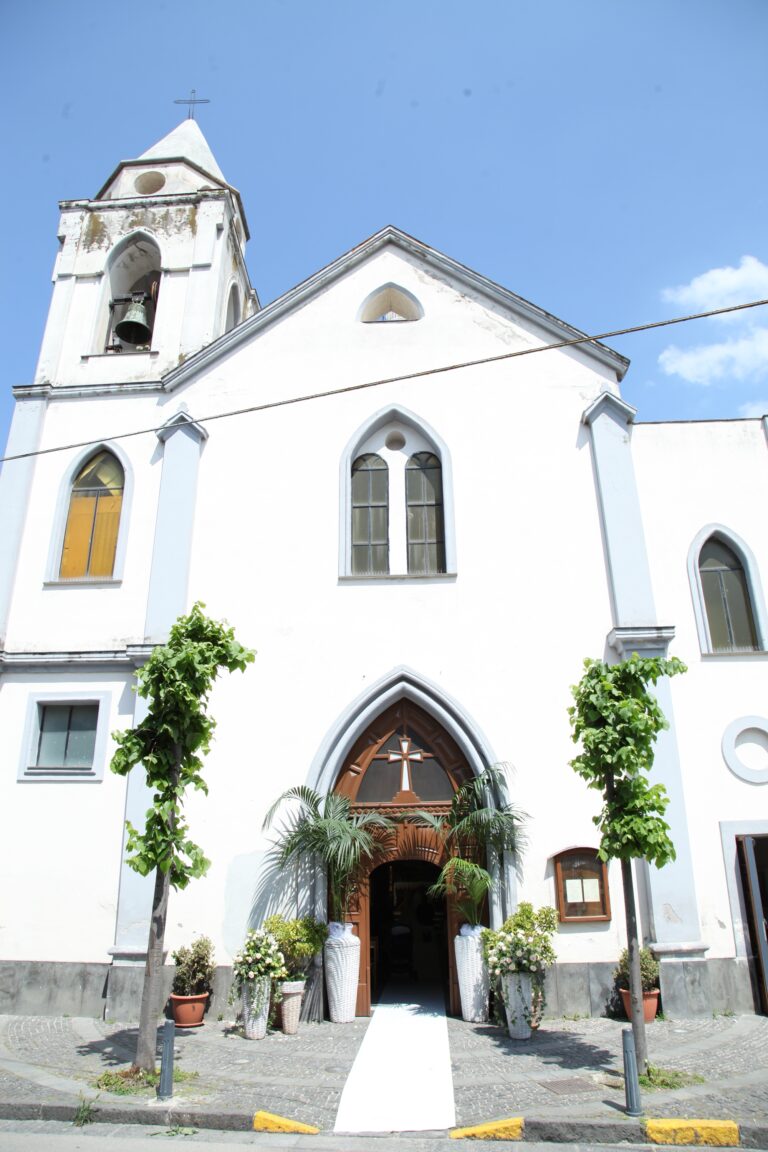 A white parish church with a bell tower, its doorway dressed with palms and a white carpet for the ceremony, WEM Eventi