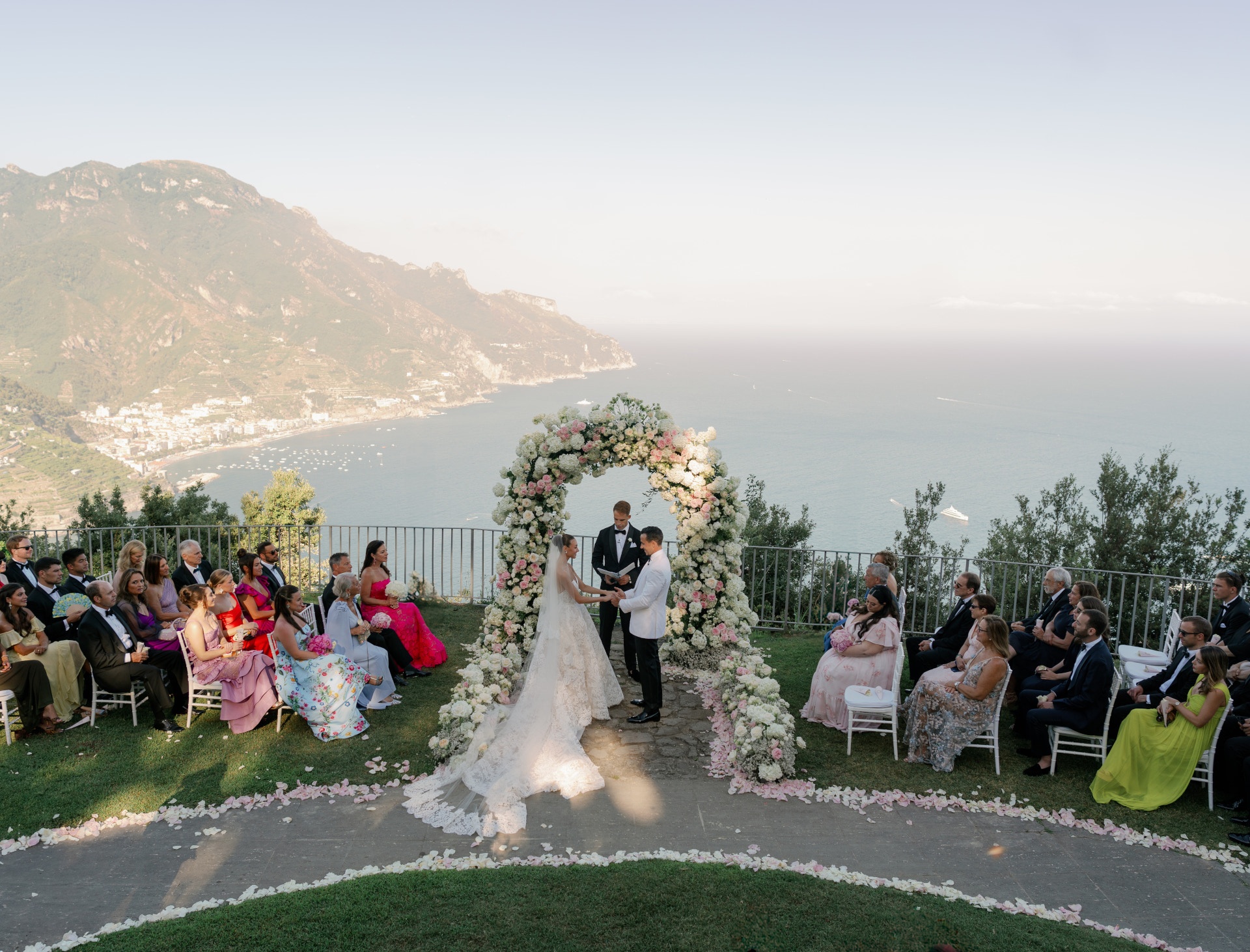 A ceremony beneath a rose and hydrangea arch, the Amalfi coastline dropping away behind the aisle, White Emotion