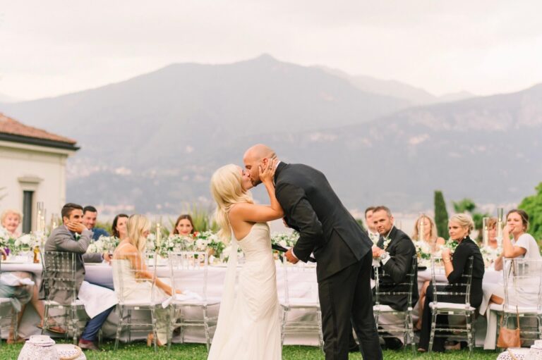 The first kiss at an outdoor reception, Lake Como's mountains behind the tables, White Emotion