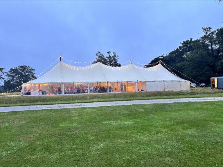 A traditional pole marquee lit with festoon lights at dusk on the marquee site, Wickham House