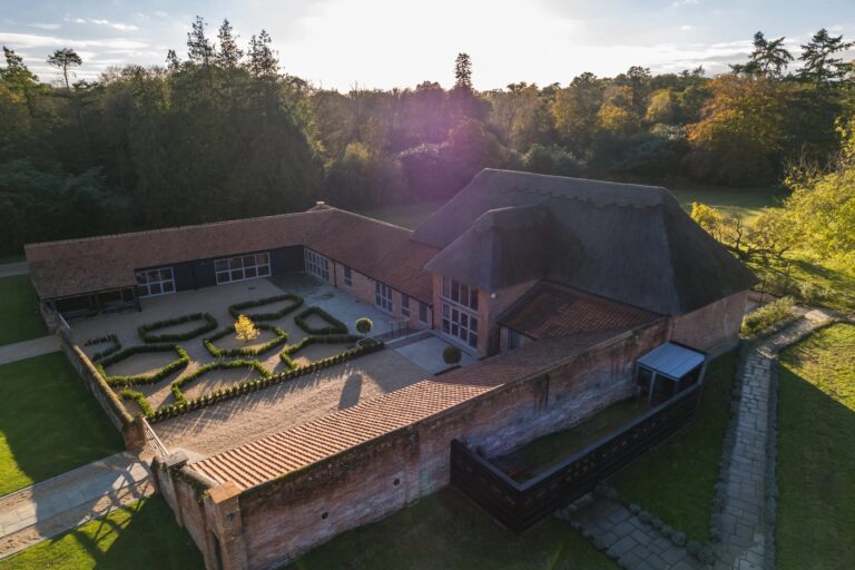 Low aerial view of The Woodyard: the thatched barn, the tiled ranges around the box-hedged courtyard and the woodland beyond, Worstead Estate