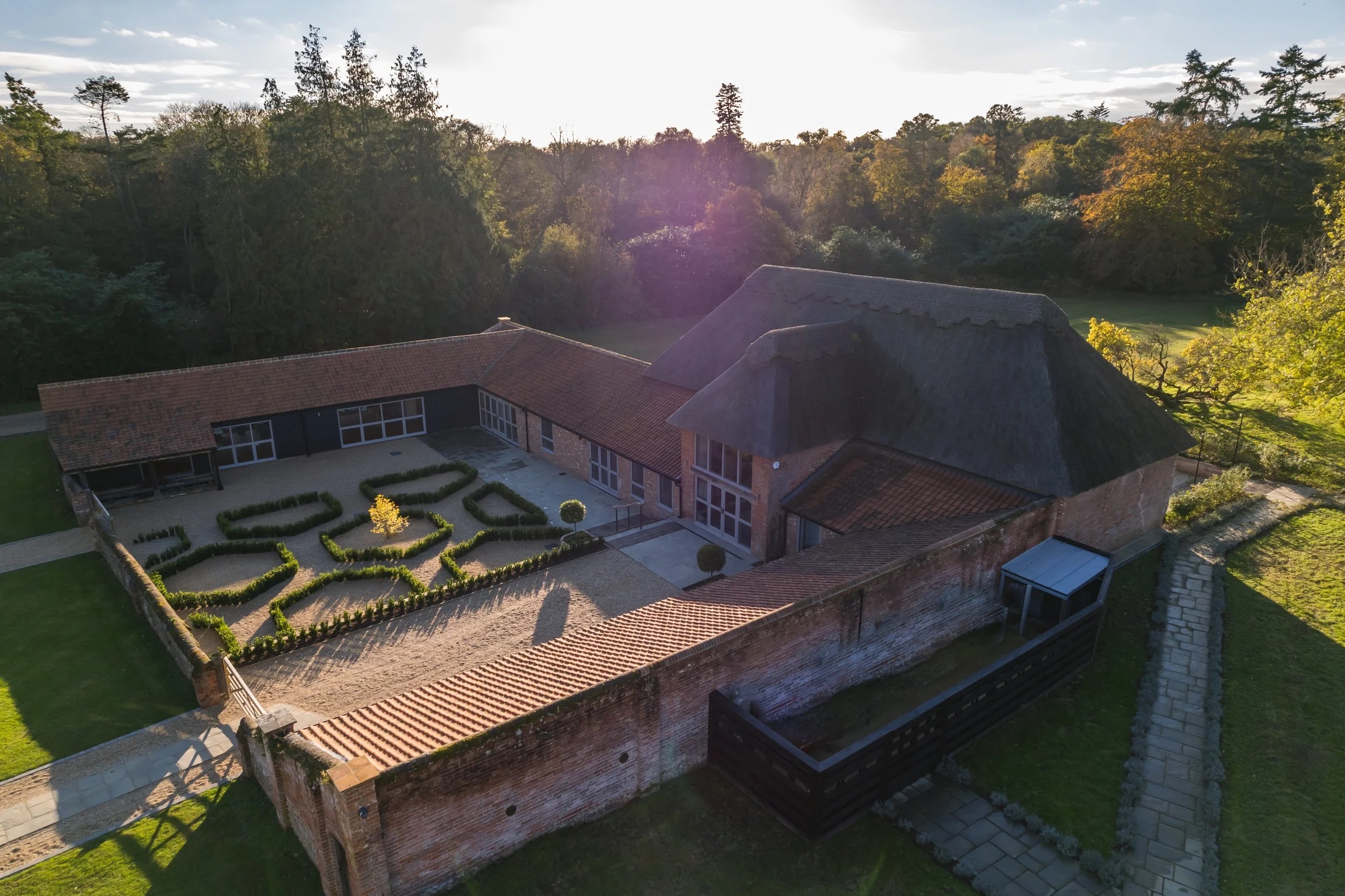 Low aerial view of The Woodyard: the thatched barn, the tiled ranges around the box-hedged courtyard and the woodland beyond, Worstead Estate