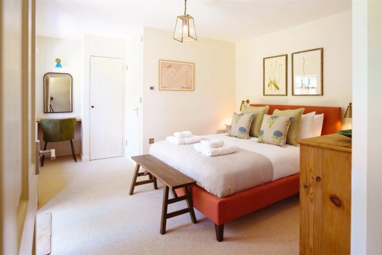 A king-size bedroom at Gatehouse North with a coral upholstered bed, a timber bench and a dressing table by the window, Worstead Estate