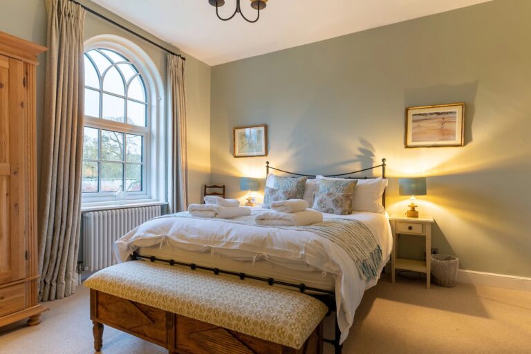 A bedroom at Estate Cottage with an arched sash window, an iron bedstead and a padded ottoman at its foot, Worstead Estate