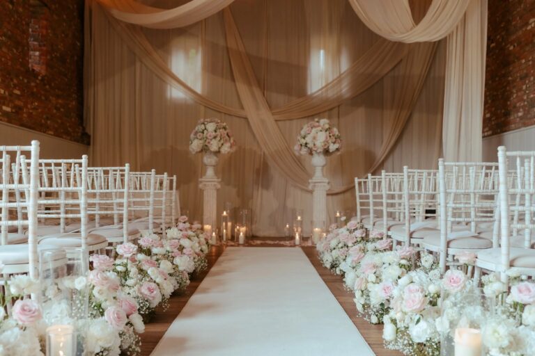 A ceremony aisle in the Thatched Barn lined with white and pale pink roses and candles, with draped fabric behind, Worstead Estate