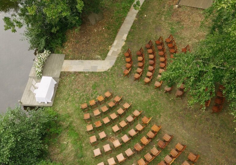 The lakeside ceremony spot from above: a wooden platform at the water, a signing table and curved rows of folding chairs on the grass, Worstead Estate