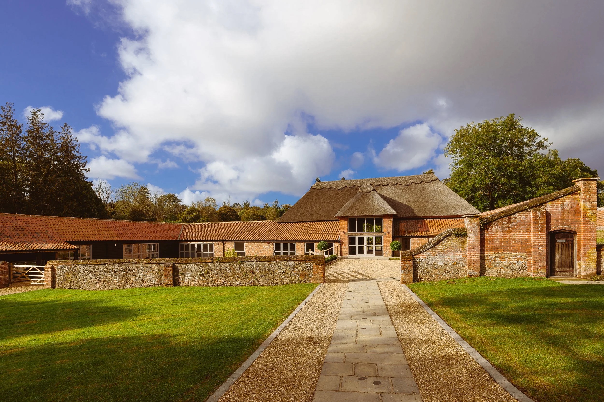 The Woodyard seen along the stone path across its front lawn, with the thatched barn rising behind the long tiled ranges and brick walls, Worstead Estate