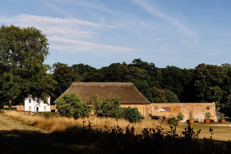 The thatched barn and walled garden seen across rough parkland grass, with the white Estate Cottage at the left and woodland behind, Worstead Estate
