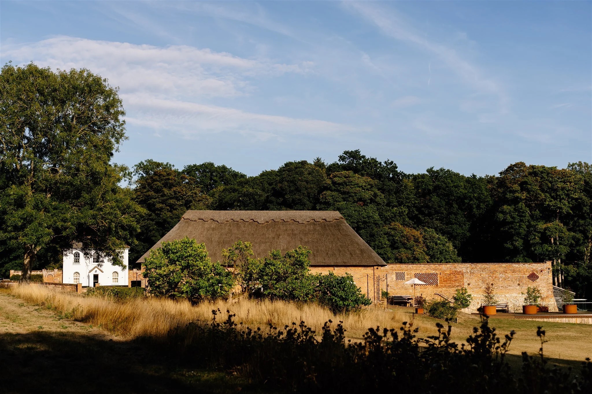 The thatched barn and walled garden seen across rough parkland grass, with the white Estate Cottage at the left and woodland behind, Worstead Estate