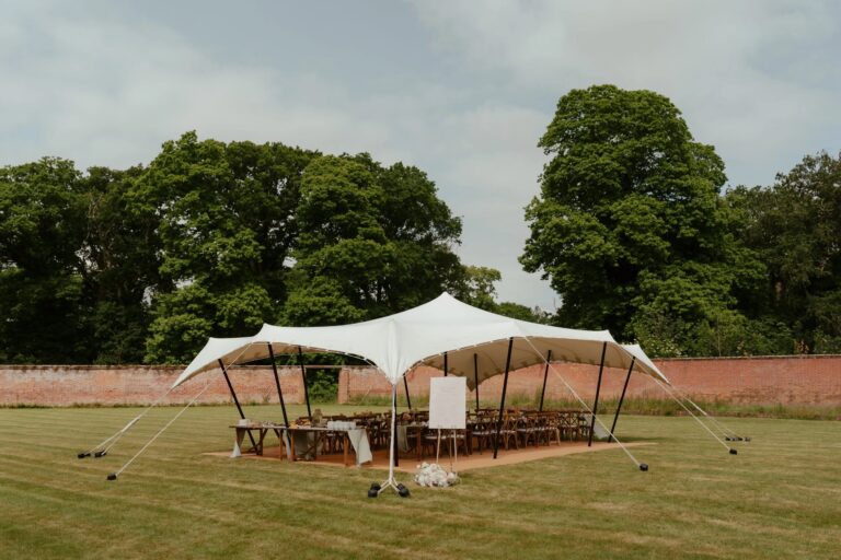 Long tables laid under a white stretch tent on the lawn of the Walled Garden, with the old brick wall and mature trees behind, Worstead Estate