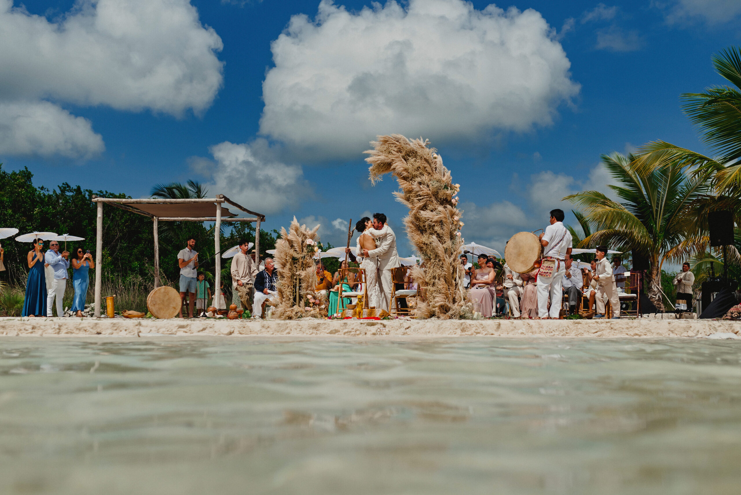 A beach ceremony set beside a sculptural driftwood arch, seen across shallow water, YD Events