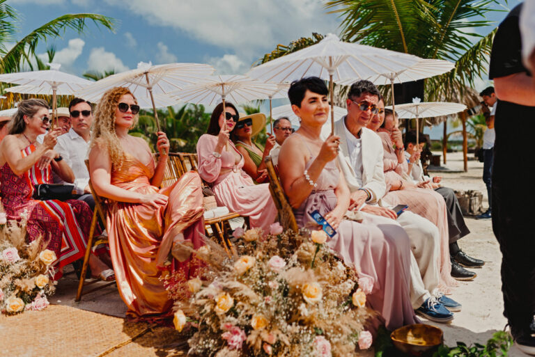 Guests in coloured outfits seated under parasols at a beach ceremony, with floral arrangements in the foreground, YD Events