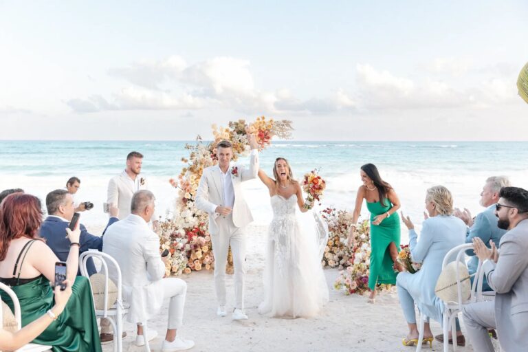 A beach ceremony beneath a floral arch with guests seated on white chairs, YD Events
