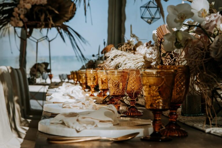 A long banquet table with amber glassware and dried florals by the sea, YD Events