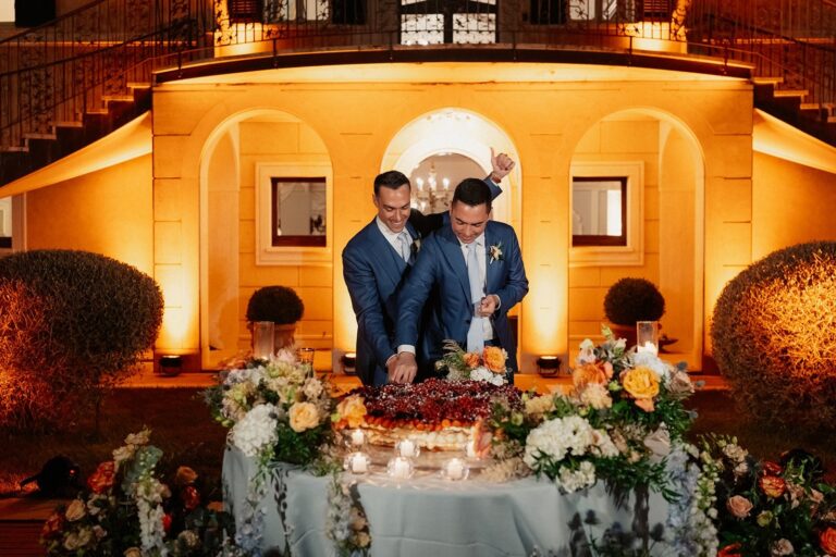 Two grooms cutting a fruit-topped cake at night under a warm-uplit stone loggia, Your Secret Tuscany